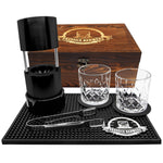 Barware set with two glasses, a black container, and tools on a black mat with a wooden box labeled 'Pressie Barware'.