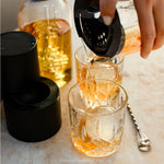Person pouring a drink into a glass using a bar spoon, with a bottle of liquor in the background. Pressice Barware
